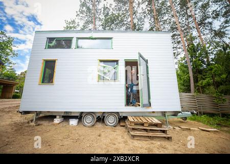 Eine junge Frau steht an der Tür eines modernen, winzigen Hauses auf Rädern, vor einem Hintergrund von Bäumen, die ein Beispiel für ein kompaktes ländliches Leben darstellen. Stockfoto