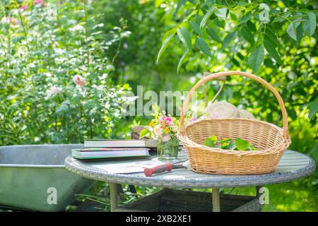 Eine ruhige Gartenszene im Freien mit Korb, Strohhut und einer Sammlung von Büchern auf einem Metalltisch, die in sanftes, natürliches Licht getaucht sind. Stockfoto