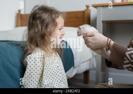 Ein junges Mädchen schaut entzückt einen Hamster an, der von den Händen ihrer abgeschnittenen, nicht erkennbaren Mutter gehalten wird, während sie zu Hause gute Zeit miteinander verbringt und ein zeigt Stockfoto