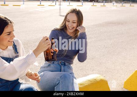 Zwei junge Frauen genießen einen sonnigen Tag im Hafenbereich, lachen und toasten mit Bierflaschen. Stockfoto