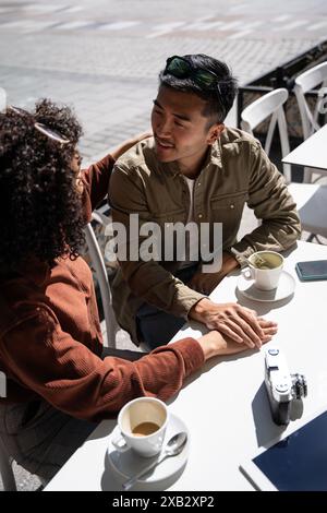 Ein chinesischer Mann und eine hispanische Frau verbringen einen intimen Moment bei einem Kaffee in einem Café im Freien in Madrid, Spanien. Sie halten die Hände und unterhalten sich, fangen sie ein Stockfoto
