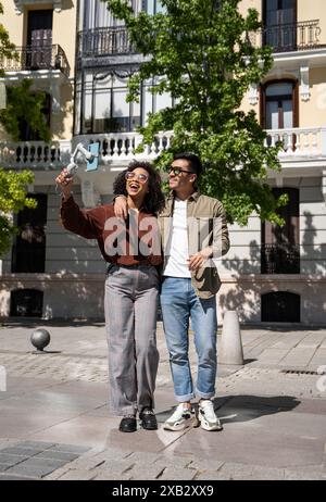 Ein fröhliches, multiethnisches Paar, ein Chinese und eine hispanische Frau, genießen einen sonnigen Tag in Madrid, Spanien. Sie machen zusammen ein Selfie vor einem Stockfoto