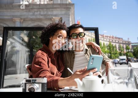 Ein multiethnisches Paar, ein Chinese und eine hispanische Frau, lächeln und umarmen sich, während sie ein Selfie mit einem Smartphone in einem Café im Freien in Madrid, Spa, machen Stockfoto