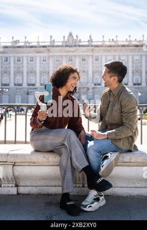 Ein multiethnisches Paar mit einem Chinesen und einer hispanischen Frau teilt einen freudigen Moment, während es vor einem historischen Gebäude in Madrid, Spa, sitzt Stockfoto