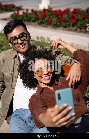 Ein fröhlicher Chinese und eine hispanische Frau, die eine stilvolle Sonnenbrille tragen, machen ein Selfie, während sie auf einer Bank in einem lebhaften Park mit roten Blumen sitzen Stockfoto