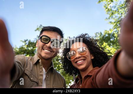 Ein multiethnisches Paar macht ein Selfie in einem sonnigen Madrid Park. Der chinesische Mann und die Hispanic-Frau lächeln, tragen eine Sonnenbrille und genießen das Outdoo Stockfoto