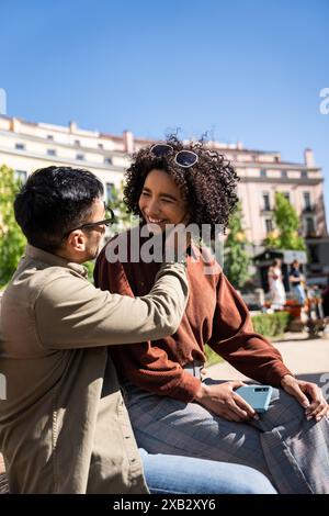 Ein fröhliches, multiethnisches Paar, ein Chinese und eine hispanische Frau, verbringen einen zärtlichen Moment, während sie draußen an einem sonnigen Tag in Madrid, Spanien, sitzen. Sie A Stockfoto