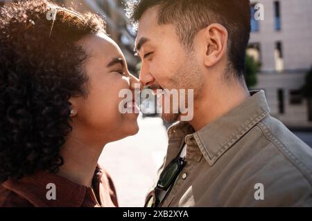 Ein liebevolles Paar, ein chinesischer Mann und eine hispanische Frau, teilen einen zarten Moment, indem sie Nasen berühren und fröhlich unter der warmen Madrider Sonne lächeln. Ihr h Stockfoto