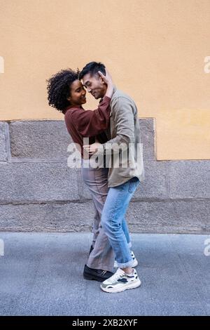 Ein multiethnisches Paar, ein chinesischer Mann und eine hispanische Frau, teilen sich eine liebevolle Umarmung an einer Stein- und gelben Mauer in Madrid, Spanien, die die Abbildung zeigt Stockfoto