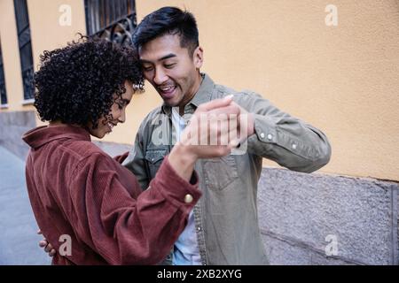 Ein fröhliches multiethnisches Paar, ein Chinese und eine hispanische Frau, tanzen zusammen auf einer bunten Straße in Madrid, Spanien. Sie teilen einen schönen Moment, EM Stockfoto