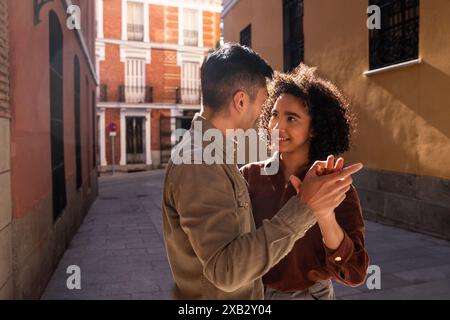 Ein chinesischer Mann und eine hispanische Frau teilen einen zärtlichen Moment auf einer sonnenbeschienenen Straße in Madrid, Spanien. Das Paar schaut sich gegenseitig in die Augen, umgeben von T Stockfoto