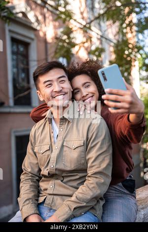 Ein multiethnisches Paar, bestehend aus einem lächelnden Chinesen und einer hispanischen Frau, umarmt sich während eines Selfie vor dem Hintergrund der urbanen Architektur Stockfoto
