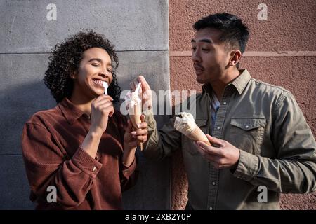 Ein fröhliches multiethnisches Paar, bestehend aus einem Chinesen und einer hispanischen Frau, die an einem sonnigen Tag in Madrid, Spanien, Eiskegel teilen. Das Paar i Stockfoto