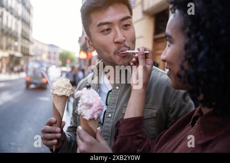 Ein multiethnisches Paar, ein chinesischer Mann und eine hispanische Frau, genießen gemeinsam Eis an einem sonnigen Tag in Madrid, Spanien. Sie teilen sich eine süße Momentfütterung Stockfoto