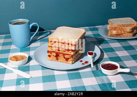 Ein Stapel von Erdnussbutter- und Jelly-Sandwiches, die gerade zubereitet wurden, mit einer Tasse amerikanischen Kaffee und einigen Brotscheiben auf blauem Hintergrund Stockfoto