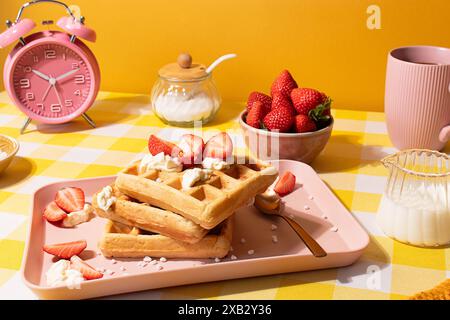 Ein lebhaftes Stillleben mit Waffeln, garniert mit Schlagsahne und frischen Erdbeeren, serviert auf einem rosafarbenen Tablett auf gelbem Hintergrund, neben einem rosafarbenen Tablett Stockfoto