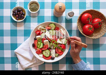 Ein frischer griechischer Salat, serviert in einer weißen Schüssel auf einer blau karierten Tischdecke, mit Tomaten, Gurken, Oliven und Fetakäse, mit einer Hand mit einem Stockfoto