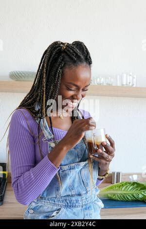 Eine fröhliche Frau mit geflochtenen Haaren lacht, während sie ein Glas Eiskaffee in gemütlicher Atmosphäre genießt und Glück und Entspannung ausdrückt Stockfoto