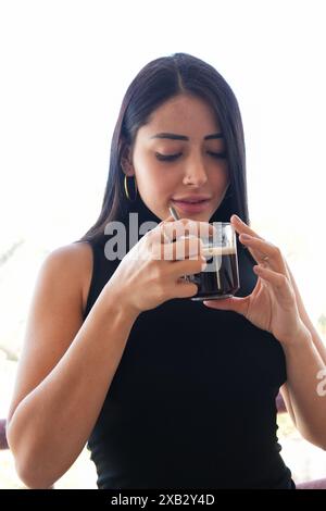 Eine junge Frau in einem schwarzen ärmellosen Oberteil, die den Geschmack von frischem schwarzem Kaffee aus einer Glasschale in einem sanft beleuchteten Innenbereich genießt Stockfoto