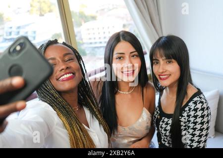 Drei junge Frauen mit unterschiedlichem Hintergrund teilen einen freudigen Moment, während sie gemeinsam ein Selfie in einem sonnendurchfluteten Raum machen. Sie zeigen ein echtes Lächeln und fas Stockfoto