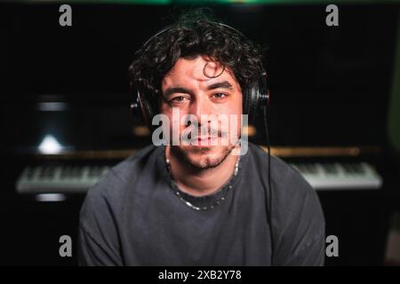 Nahaufnahme eines jungen männlichen Musikers, der Kopfhörer in einem Studio trägt, mit einem unscharfen Klavier im Hintergrund, was seine kreative Umgebung unterstreicht Stockfoto