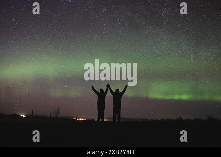 Silhouetten von zwei Individuen mit erhobenen Armen unter einer lebhaften Darstellung der Nordlichter am isländischen Nachthimmel, die die atemberaubende Natur zeigen Stockfoto