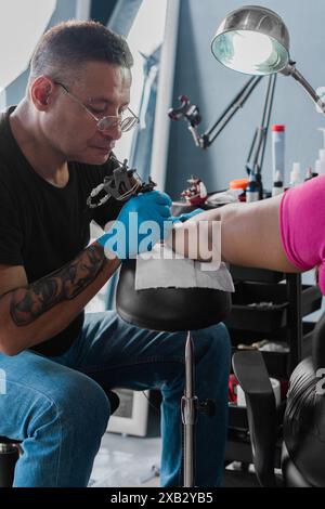 Ein fokussierter hispanischer Tätowierer färbt geschickt ein Design auf den Arm seines Kunden in einem gut ausgestatteten Studio in Ecuador. Er blickt auf seinen Wor herunter Stockfoto