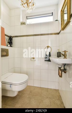 Elegantes klassisches Badezimmer mit weißer Keramik-Toilette, Sockelwaschbecken, gefliesten Wänden mit goldener Zierleiste und luxuriösem Kronleuchter in Ros Stockfoto