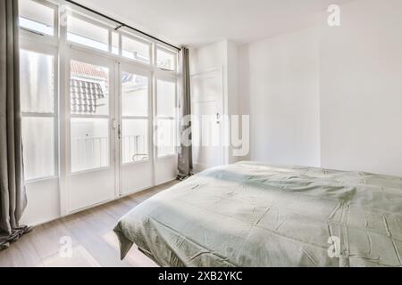 Ein gut beleuchtetes, modernes Schlafzimmer mit großen französischen Fenstern, einem komfortablen Bett mit pastellfarbener Tagesdecke und minimalistischen weißen Wänden, die eine ruhige Atmosphäre schaffen Stockfoto