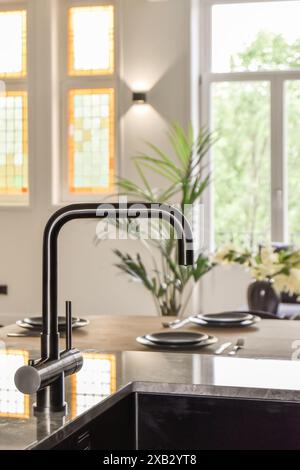 Aus nächster Nähe sehen Sie den schwarzen Wasserhahn und das Waschbecken einer modernen Küche mit verschwommenem Hintergrund, einem Esstisch, lebendigen Buntglasfenstern und einem Innern Stockfoto