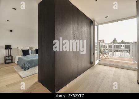 Dieses Bild zeigt ein modernes Schlafzimmer mit einem großen Bett mit frischer Bettwäsche, einer dunklen Holzwand und einer offenen Terrassentür, die zu einem Balkon führt Stockfoto