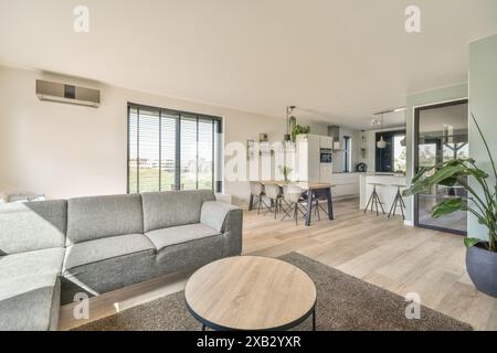 Ein modernes Wohnzimmer fügt sich nahtlos in einen stilvollen Küchenbereich mit großen Fenstern, hellem Holzfußboden und modernen Möbeln ein Stockfoto