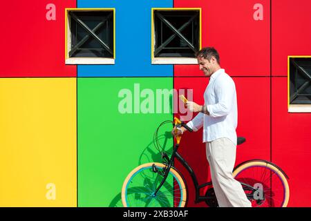 Ein stilvoller Mann, der sich auf ein farbenfrohes Fahrrad stützt, während er sein Smartphone vor einer lebendigen, mehrfarbigen Wand mit geometrischen Formen liest Stockfoto