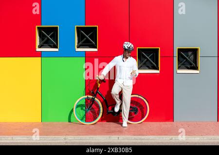 Ein Mann in weißer Kleidung lehnt sich auf ein farbenfrohes Fahrrad vor eine bunte, bunte Wand, während er auf sein Handy blickt Stockfoto