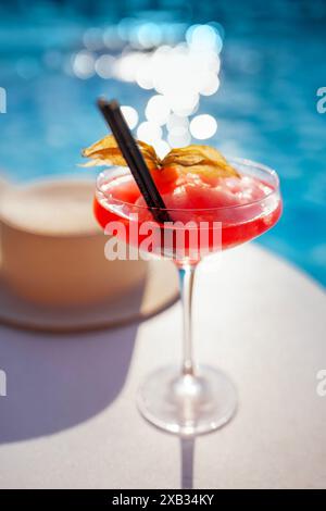 Ein rotes Getränk in einem eleganten Glas mit langem Stiel an der Seite des Pools. Ein alkoholisches Eisgetränk, dekoriert mit einer Blume auf einem Hintergrund aus Türkis Stockfoto