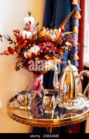 Ein schöner runder Glastisch mit einem Wasserkocher aus Metall, einem Milchkännchen und einer Tasse. Herbstlicher Blumenstrauß mit Baumwolle und eleganten Gerichten. Details zu A Stockfoto