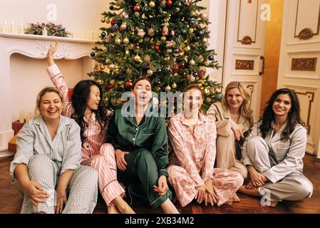 Junge Frauen im Pyjama lachen und sitzen unter einem geschmückten Weihnachtsbaum im Wohnzimmer. Lächelnde Multi-Rassen-Mädchen auf einen Weihnachts-Vater Stockfoto