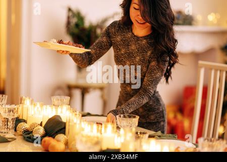 Ein attraktives junges asiatisches Mädchen stellt einen festlichen Tisch mit Neujahrsdekoration und beleuchteten Kerzen. Eine charmante lächelnde Frau in einem eleganten Kleid hält einen Stockfoto