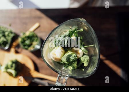 Nahaufnahme von grünem Gemüse, Banane und Kräutern in einem Mixer. Welliger Kohl, Brokkoli und Rucoli auf einem Holztisch. Draufsicht. Herstellung eines Vitamins Stockfoto