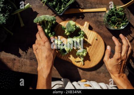 Weibliche Hände zerlegt in Brokkoli-Blütenstände auf Holztisch. Draufsicht. Die junge Frau bereitet vitaminreiche grüne Salate aus Rucola und Cabb zu Stockfoto