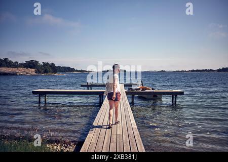 Blick auf die gesamte Länge von hinten auf eine lächelnde Frau, die an sonnigen Tagen auf dem Steg über dem See spaziert Stockfoto