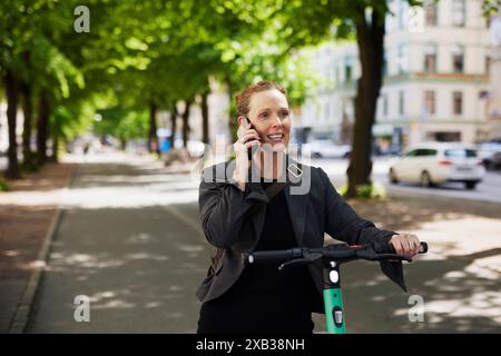 Lächelnde Geschäftsfrau mit elektrischem Roller, die auf dem Fußweg über das Smartphone spricht Stockfoto