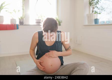 Schwangere junge Frau, die auf den Bauch blickt, während sie auf der Matte sitzt Stockfoto