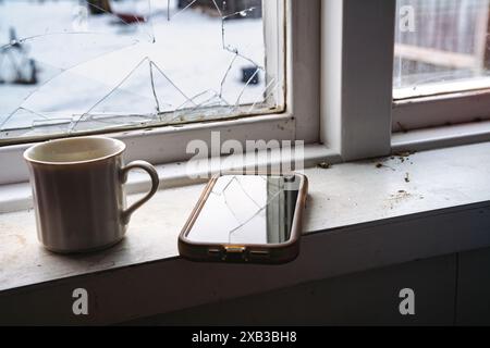 Nahaufnahme des Smartphones und der Kaffeetasse durch kaputtes Fenster Stockfoto