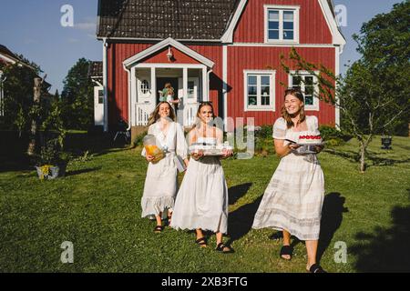 Porträt in voller Länge von glücklichen jungen Freundinnen, die an sonnigen Tagen Essen und Getränke im Hinterhof tragen Stockfoto