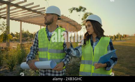 Kaukasische Kollegen Architektenpartnerschaft männlicher Mann Geschäftsmann Ingenieur halten Blueprint, der Talk erklärt weibliche Geschäftsfrau Inspektorin Frau schreibt Stockfoto