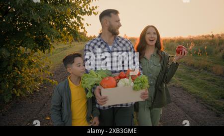 Glückliche kaukasische Familie Rückkehr nach der Landwirtschaft Arbeit ernten Mann männlicher Kerl Vater Tragen Kiste Gemüse kleines Kind Sohn Junge spazieren Frau Stockfoto