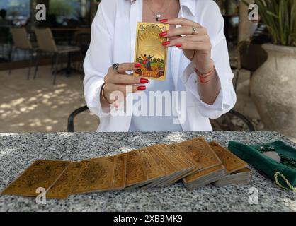 Montenegro, Budva, 6. Mai 2024. Eine Frau in einer weißen Bluse mit roten Nägeln hält die Ten of Cups Tarot-Karte, während andere Tarot-Karten auf einem Tisch liegen. Fahrer-Waite Tarot Stockfoto