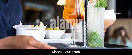 Der Kellner serviert erfrischende Cocktails und Snacks auf einem Tablett in einer stilvollen Bar. Die Getränke beinhalten einen Orangenspritz und einen Pfefferminz garnierten Cocktail, perfekt Stockfoto