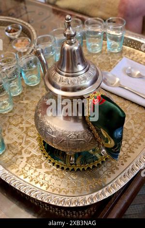 Polierte marokkanische Zeremonie-Teekanne mit traditioneller Dekoration, Griffabdeckung, Teekannenständer und Gläsern, die auf einem versierten silbernen Serviertablett stehen. Stockfoto